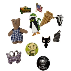 Könnte beinhalten: Eine Sammlung verschiedener Anstecknadeln, darunter ein brauner Teddybär in blau karierter Latzhose, ein Colorado-Pin, eine US-Flagge, ein Pinguin mit Zuckerstange, eine Panflöte, ein Stinktier, eine schwarze Katze, ein Wolf und ein lila Schmetterling.