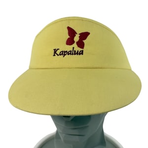 May include: A pale yellow visor featuring an embroidered dark red butterfly icon positioned above the text Kapalua in a matching dark red font. The visor has a wide, curved brim and a structured crown, providing a simple and functional design.