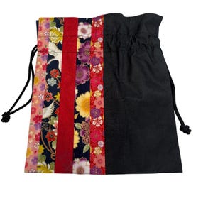 May include: A drawstring pouch featuring a vertical patchwork design on one side with floral and crane patterns in pink, red, yellow, and dark blue, contrasted by a solid black fabric panel on the other side with black cord drawstrings.