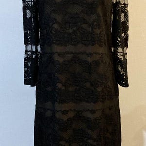 Könnte beinhalten: Ein schwarzes Spitzenkleid mit langen Ärmeln und einem U-Boot-Ausschnitt. Das Kleid besteht aus zarter Spitze mit floralem Muster. Das knielange Kleid hat eine figurbetonte Silhouette, perfekt für eine formelle Veranstaltung oder einen besonderen Anlass.