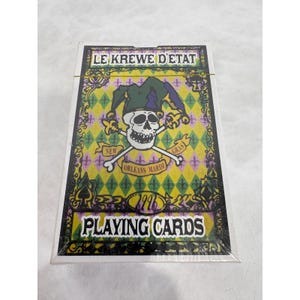May include: A sealed deck of playing cards with a colorful design. The card box features a skull wearing a jester's hat, crossed bones, and the text "LE KREWE D'ETAT" and "PLAYING CARDS". The design includes fleur-de-lis and diamond patterns.