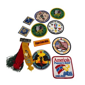 May include: A collection of embroidered scouting patches and a Webelos pin with red and yellow knit fabric tails. The patches feature various designs including a bobcat, an eagle, a sunset, and text such as Webelos Weekend, Tiger Cubs BSA, and America's Popcorn Sale.