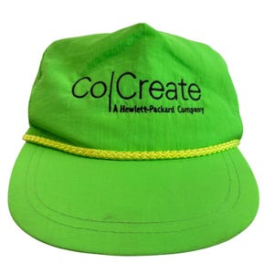 May include: A neon green baseball cap featuring a yellow braided cord trim across the base of the crown. The front panel displays the black embroidered text "co|Create A Hewlett-Packard Company" in a clean, sans-serif font style.