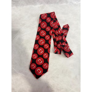 May include: A black necktie with a repeating pattern of red circles, each containing a white letter "C". The tie is made of a smooth fabric and is displayed on a white surface. The tie is a classic accessory.