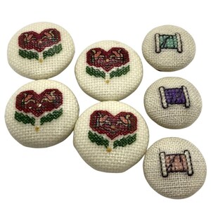 May include: A set of seven round fabric-covered buttons featuring cross-stitch embroidery. Four buttons display a red heart-shaped floral design with green leaves, while three buttons show a spool of thread in light green, purple, and pink.