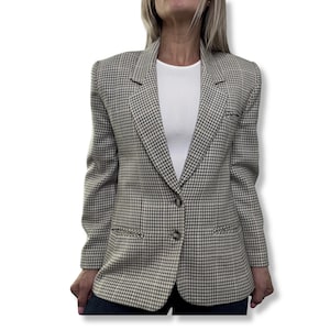 Könnte beinhalten: Ein Blazer mit Hahnentrittmuster in Grau- und Beigetönen. Der Blazer hat ein klassisches Revers, zwei Vordertaschen und einen Zwei-Knopf-Verschluss. Die Jacke wird über einem weißen Oberteil getragen.