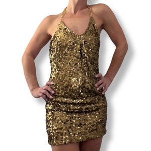 Op de afbeelding: Een gouden halterjurk met pailletten, een V-hals en een kettingband. De jurk is mini-lengte en bedekt met glinsterende pailletten. De jurk is effen goudkleurig.