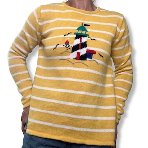 Può includere: Maglione a righe gialle e bianche con un disegno di faro. Il faro è nero e bianco con accenti rossi e blu, su una spiaggia sabbiosa con gabbiani. Il maglione ha maniche lunghe.