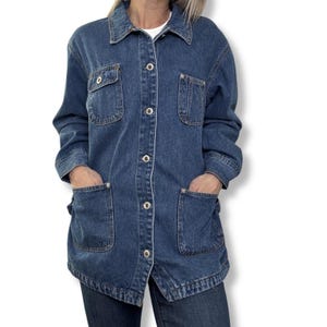 May include: A mid-wash denim jacket with a collared neckline, button-up front, and four pockets. The jacket has a relaxed fit and is worn over a white shirt. The sleeves are long, and the jacket falls to mid-thigh. The jacket is made of a sturdy denim material.