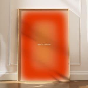 Könnte beinhalten: Gerahmter Kunstdruck mit einem leuchtenden orange-gelben Farbverlauf. Der weiße Text lautet "good to be home". Das Kunstwerk wird in einem goldenen Rahmen vor einer weißen Wand und einem Holzboden präsentiert.