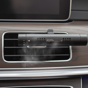 Puede incluir: Un ambientador de coche negro está montado en una rejilla de ventilación del coche. El dispositivo cilíndrico tiene una serie de pequeños agujeros y un diseño en escritura. Se ve una voluta de vapor. El interior del coche tiene un acabado de vetas de madera.