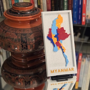 May include: An ornate, tiered, orange and black lacquered container with detailed patterns. Next to it is a white framed map of Myanmar, with the country's name in orange. The map is multi-coloured.