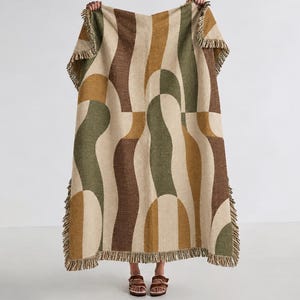May include: A rectangular woven throw blanket featuring a repeating abstract pattern of curved, interlocking shapes in shades of olive green, mustard yellow, and dark brown against a cream background, finished with a fringed edge on all sides.