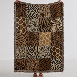 May include: A rectangular woven throw blanket featuring a patchwork design of various animal prints including leopard spots, zebra stripes, and giraffe patterns in shades of brown, black, tan, and cream, finished with a decorative fringed edge.
