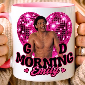 May include: A white ceramic mug with a pink handle and rim, featuring a graphic of a shirtless man centered inside a large pink disco ball heart. Below the heart, the text reads "GOOD MORNING Emily" in bold pink letters with small pink hearts.