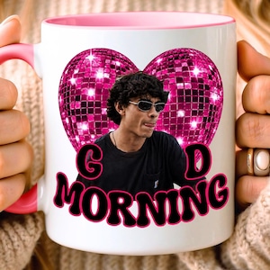 May include: A white ceramic mug with a pink handle and interior rim. The front features a graphic of a person wearing sunglasses in front of a large pink disco ball heart. Below the image, the text "GOOD MORNING" is printed in black letters.
