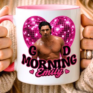 May include: A white ceramic mug with a pink interior and handle. The front features a graphic of a shirtless man centered between two pink disco ball hearts. Below the image, the text reads "G D MORNING Emily" in black and pink letters.