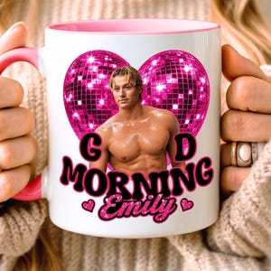 May include: A white ceramic mug with a pink handle and interior rim. The front features a graphic of a shirtless person in front of two pink glittery disco balls, with the black text "GOOD MORNING Emily" and two small pink hearts below.