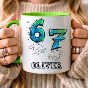 May include: White ceramic mug with a green handle and interior. The mug features the numbers "67" in blue and green, with a splatter design, and the name "OLIVER" in black block letters.