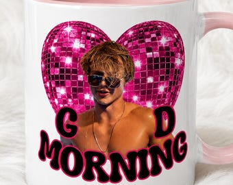 Puede incluir: Una taza de cerámica blanca con interior y asa de color rosa claro. El frente presenta un gráfico de una persona con gafas de sol frente a un gran corazón de bola de discoteca rosa. Debajo, el texto GOOD MORNING en letras negras.
