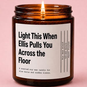 May include: A lit soy wax candle in a brown glass jar with a white label. The text reads: Light This When Ellis Pulls You Across the Floor, Soy Wax Blend, 50-60 Hours, Hand Poured, A scented soy wax candle for slow burns and sudden kisses.