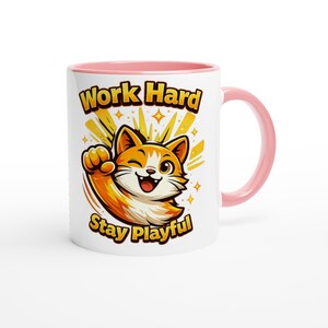 Puede incluir: Taza de cerámica blanca con interior y asa rosa. La taza presenta una ilustración de gato de dibujos animados con el texto "Work Hard Stay Playful" en dorado y negro.