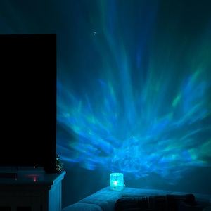 Puede incluir: Un pequeño proyector de luz cilíndrico turquesa está sobre un sofá, proyectando un vibrante efecto de aurora boreal azul y verde en la pared. La habitación está tenuemente iluminada, con un televisor oscuro y un armario blanco visibles.