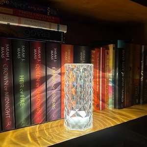 May include: A clear, faceted crystal lamp with a geometric design sits on a wooden shelf. The lamp is illuminated from within, casting a soft glow. Books with colorful spines are visible in the background.