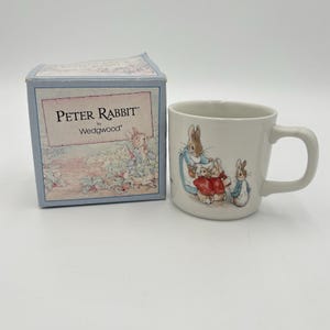 May include: A white ceramic mug featuring a colorful illustration of Peter Rabbit and family, accompanied by a matching square cardboard box labeled Peter Rabbit by Wedgwood with a garden scene printed on the front panel.