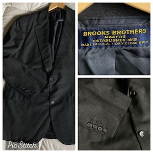 May include: A dark gray Brooks Brothers blazer with a classic three-button closure and buttoned cuffs. The interior lining is visible, with a label that reads "Brooks Brothers Makers, Established 1818, Made in U.S.A., Dry Clean Only."