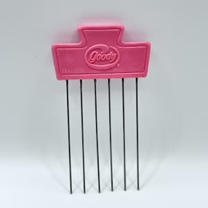 May include: A vintage pink Goody hair pick with six black metal teeth. The pink plastic top features the Goody logo and the text "U.S.A." in white. The pick is designed for styling and detangling hair.
