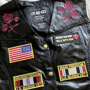 May include: Black leather vest with embroidered pink roses and patches. Patches include the American flag, "EXERCISE YOUR HEART WALK WITH GOD", "ENDURING FREEDOM U.S. MARINE VET", and "IRAQI FREEDOM U.S. MARINE VET". The vest has the text "LIVE-RIDE-ROCK BUFFALO LEATHER".