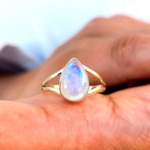 May include: A silver ring featuring a pear-shaped moonstone cabochon set in a smooth bezel. The band is split into two thin, polished silver wires that meet at the base of the stone, creating an open, airy design with a metallic finish.
