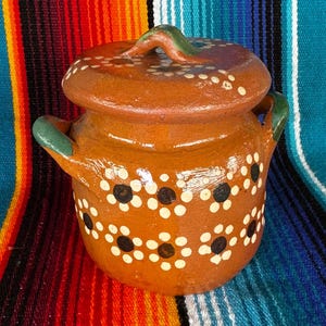 May include: A small, round, terracotta-colored ceramic pot with a matching lid. The pot features a floral pattern of white dots with black centers. The handles and lid handle are green. The pot is displayed on a colorful striped textile.