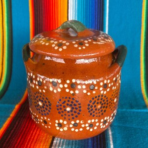 May include: A small, round, terracotta-colored ceramic pot with a lid. The pot is decorated with white and blue dots. It has two small handles and a green handle on the lid. The pot is sitting on a colorful striped textile.