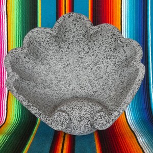 May include: A gray, speckled stone molcajete, a traditional Mexican mortar and pestle. The bowl has a scalloped edge and sits on a colorful striped textile background. This kitchenware is used for grinding spices and making salsa.