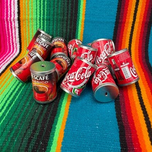 May include: A collection of small metal cans featuring red and white branding for Nescafé Clásico coffee and Coca-Cola. The cans are cylindrical with silver metal lids, some showing a small hole in the center. The Coca-Cola cans display the iconic white script logo on a red background, while the Nescafé cans feature a graphic of a red mug filled with coffee.