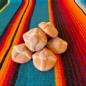 May include: A small pile of five round, golden-brown baked bread rolls, each featuring a distinct cross-shaped dough decoration on top and a light dusting of sugar, arranged in a cluster to showcase their textured, baked crusts and rounded shapes.