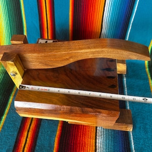 Op de afbeelding: Een handgemaakte houten tortillapers, met een donkere houten afwerking. De pers heeft een gebogen handvat en meet ongeveer 28 cm in lengte. De achtergrond is een kleurrijk gestreept textiel.