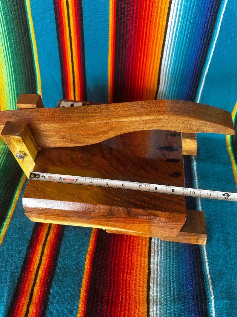 Op de afbeelding: Een handgemaakte houten tortillapers, met een donkere houten afwerking. De pers heeft een gebogen handvat en meet ongeveer 28 cm in lengte. De achtergrond is een kleurrijk gestreept textiel.