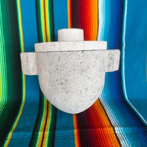 May include: A gray stone mortar and pestle set with a lid. The mortar has two side handles and a rounded bottom. The background is a colorful striped textile with blue, red, orange, and green stripes.