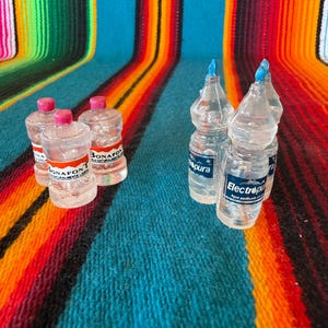 May include: A collection of miniature clear plastic water bottles featuring two distinct brands. Two bottles display the Bonafont label with pink caps, while two bottles feature the Electropura label with blue caps, all arranged in two small groups.