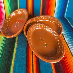 May include: Four oval-shaped, brown ceramic serving dishes with a decorative green and white dotted border. The dishes are arranged on a colorful striped textile background, showcasing a rustic, handcrafted aesthetic.