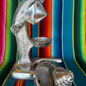 May include: A vintage, silver-colored manual citrus juicer with the letters "JMSA" visible. The juicer is made of metal and has a lever-operated design. The juicer is shown with its parts separated. The background is a colorful striped textile.