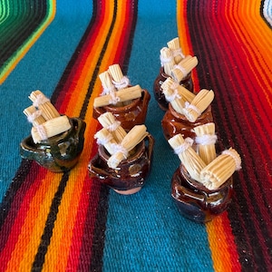 May include: A collection of six small, dark brown and green glazed ceramic pots, each containing miniature bundles of light tan corn husks tied with white string. The pots are arranged in a cluster, showcasing their glossy, rounded forms.