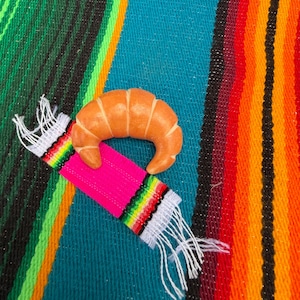 May include: A golden-brown, crescent-shaped croissant pastry resting on a small, rectangular woven fabric strip featuring a bright pink center, multi-colored striped borders in green, yellow, and red, and white fringed ends on both sides.