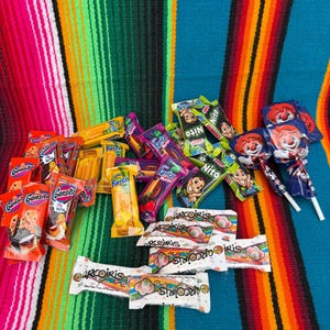 May include: A collection of assorted individually wrapped snacks and candies including Gansito, Barritas, Nito, and Arcoiris treats, along with two lollipops featuring a clown graphic, all arranged together in a group on a colorful striped surface.