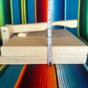 Puede incluir: Una prensa de tortillas de madera clara con mango y tapa con bisagras. La prensa tiene aproximadamente 5 cm de grosor. El fondo presenta un textil a rayas colorido.