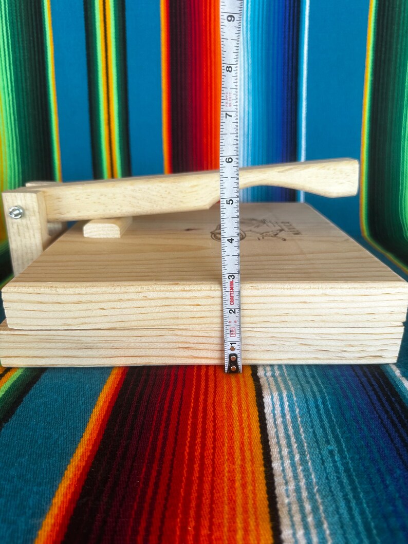 Puede incluir: Una prensa de tortillas de madera clara con mango y tapa con bisagras. La prensa tiene aproximadamente 5 cm de grosor. El fondo presenta un textil a rayas colorido.