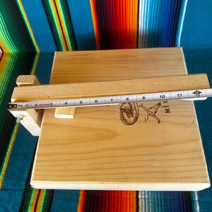 Puede incluir: Prensa de tortillas de madera clara con tapa con bisagras y asa. La prensa tiene un logotipo estampado en la superficie. Una cinta m&eacute;trica muestra una longitud de aproximadamente 30 cm.
